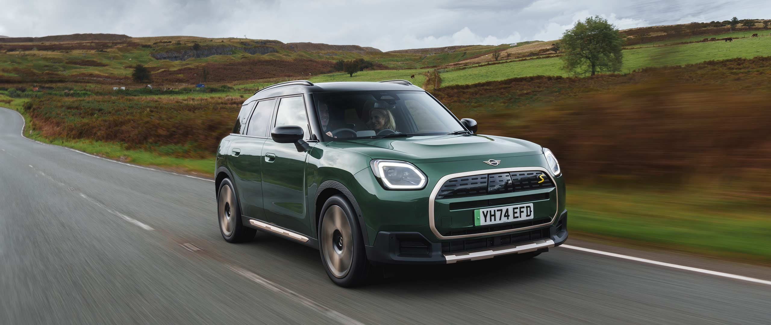 Side view of a driving all-electric MINI Countryman in Smokey Green with Vibrant Silver Roof off-road.